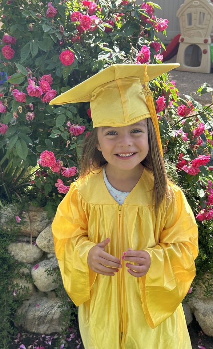 Smiling preschool graduate in a yellow cap and gown by the rose garden