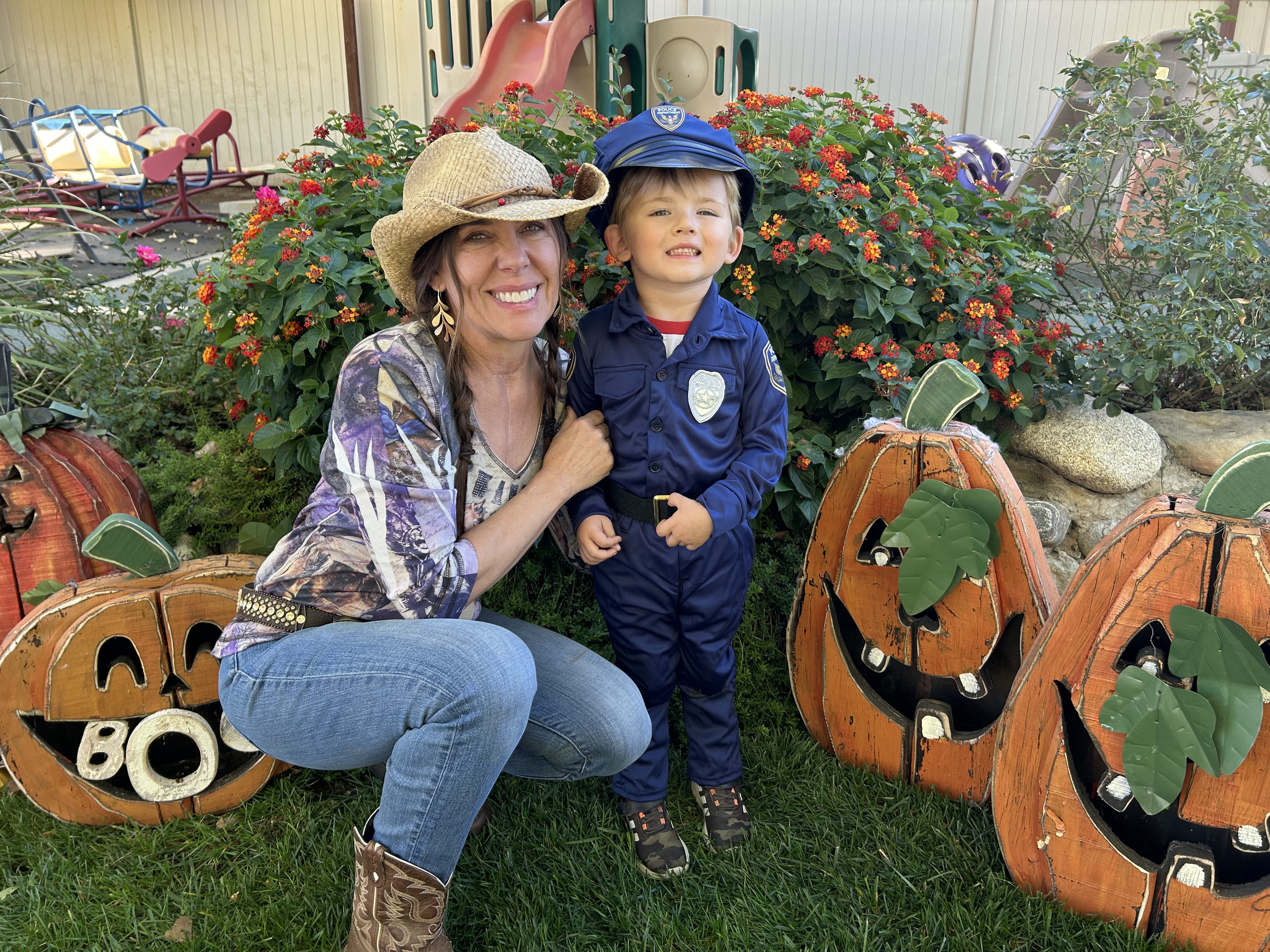 Miss Robin in a cowboy hat with a preschooler dressed as a police officer beside wooden jack-o'-lanterns in the backyard