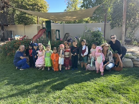 Miss Robin and the preschoolers in Halloween costumes posing together on the backyard lawn