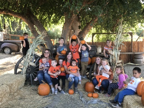 Miss Robin and the preschoolers in orange shirts posing with pumpkins under a big tree at the pumpkin patch