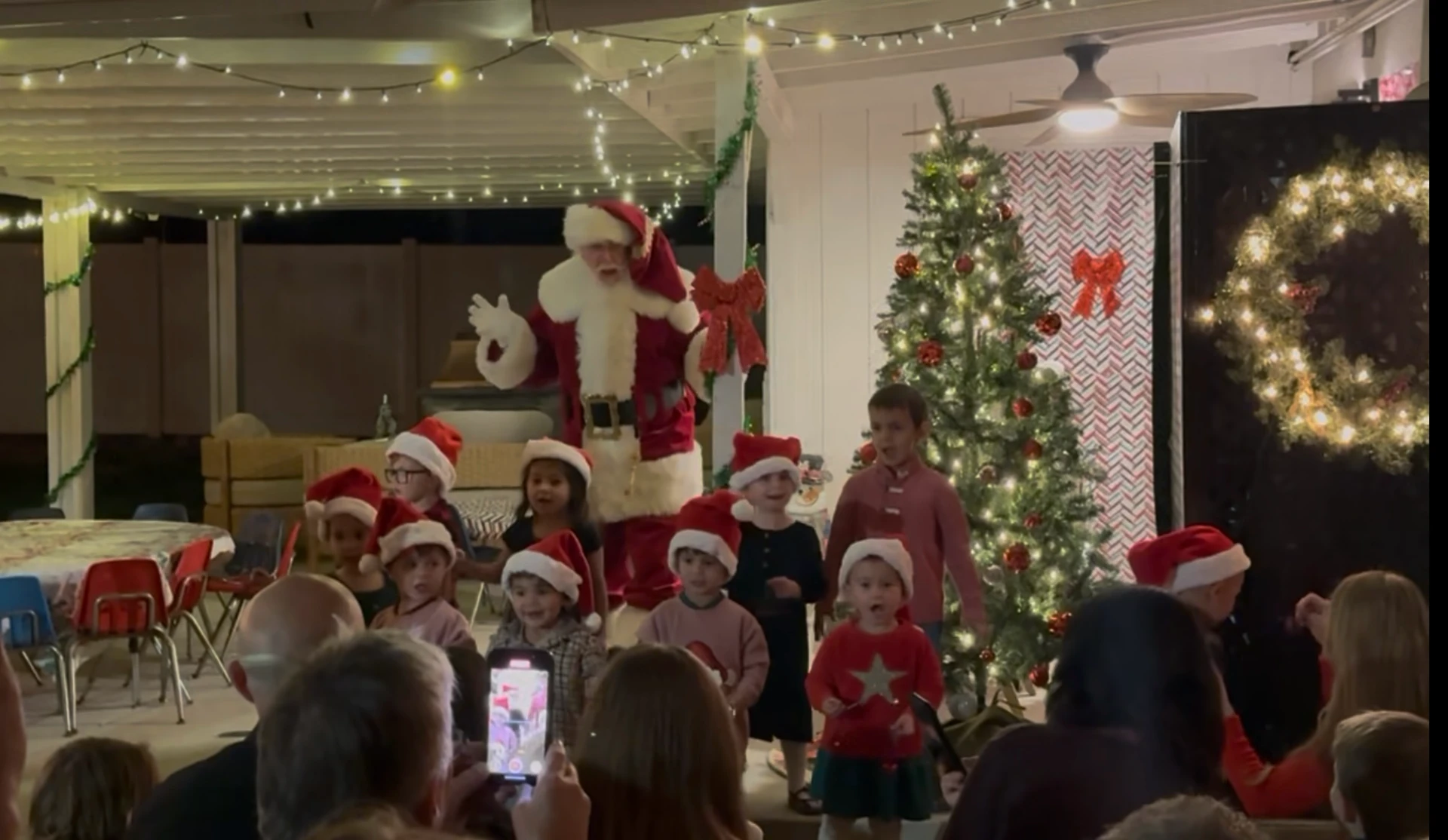 Santa and preschoolers in Santa hats singing carols beside the Christmas tree on the patio