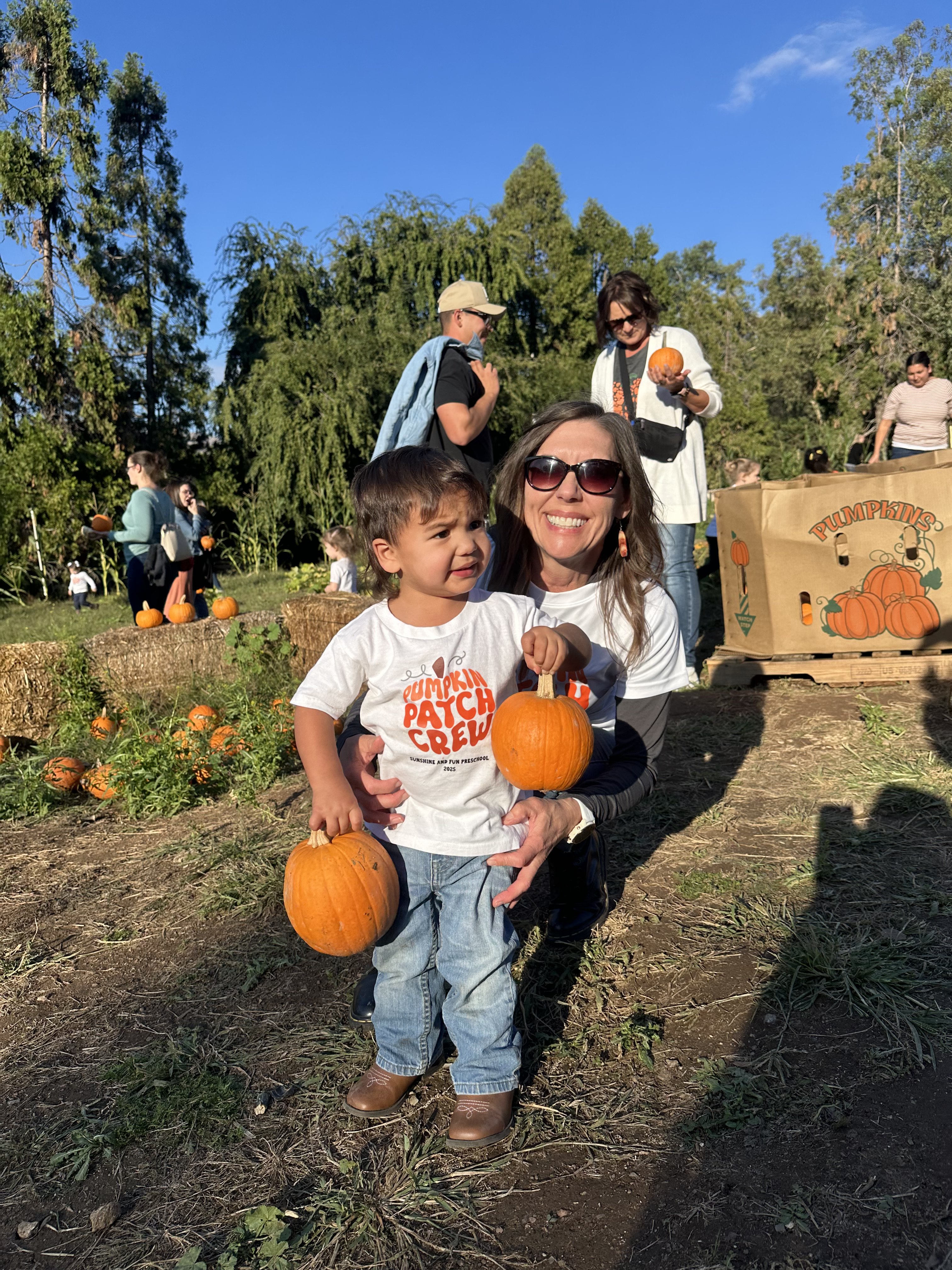 Miss Robin holding a preschooler carrying two pumpkins at the pumpkin patch