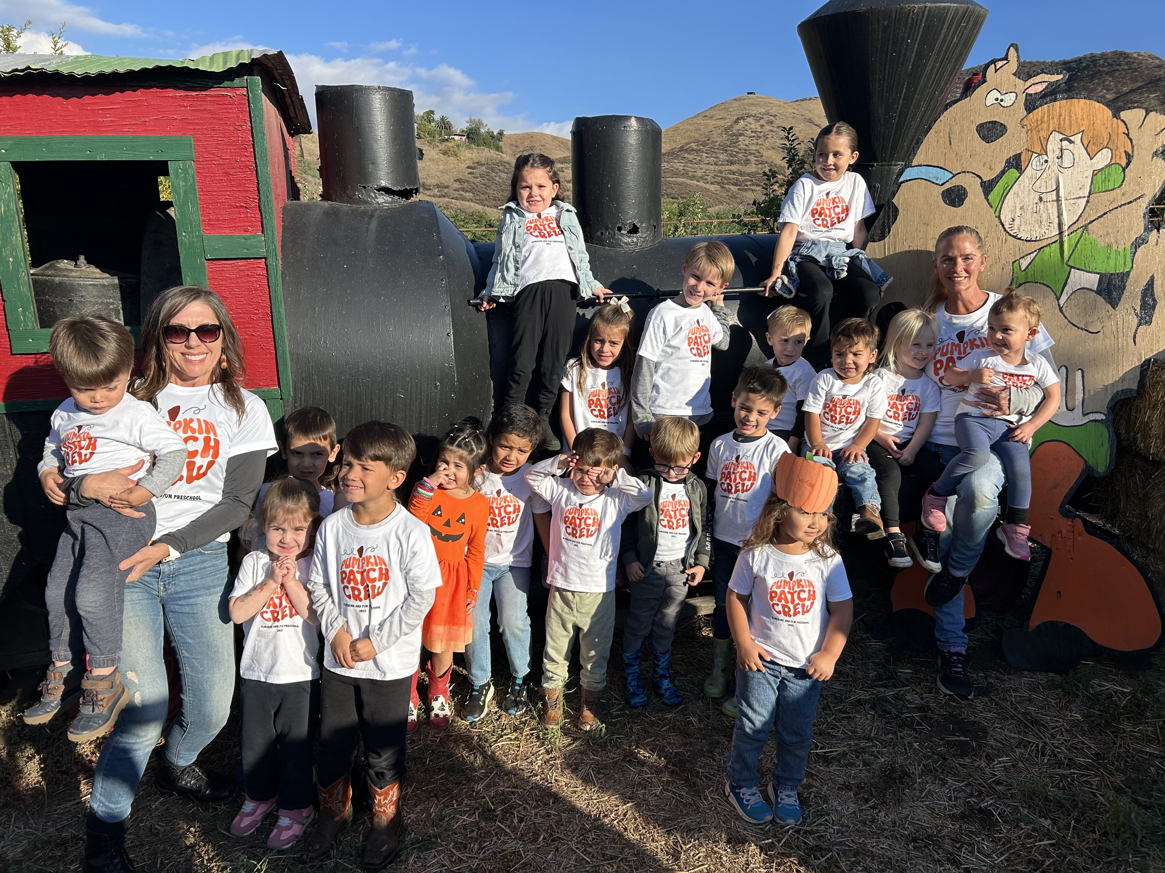 Miss Robin and the preschoolers in Pumpkin Patch Crew shirts posing by the train at the pumpkin patch