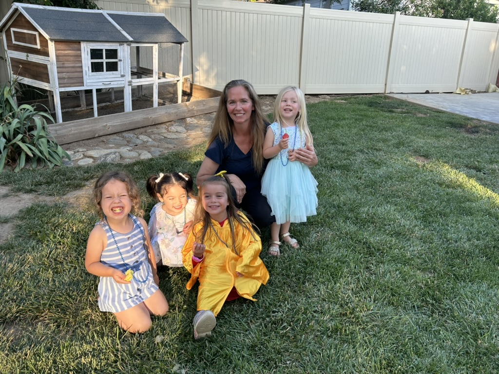 Miss Robin kneeling with smiling preschoolers on the backyard lawn, one in a gold graduation gown