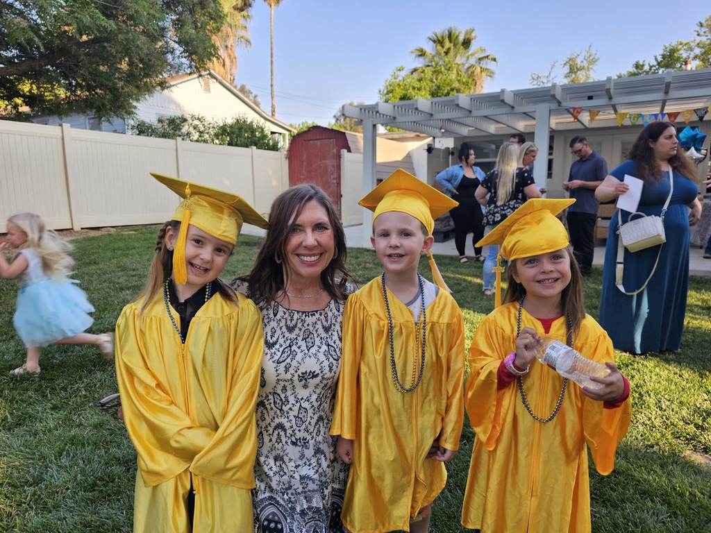 Miss Robin smiling with three preschool graduates in yellow caps and gowns in the backyard