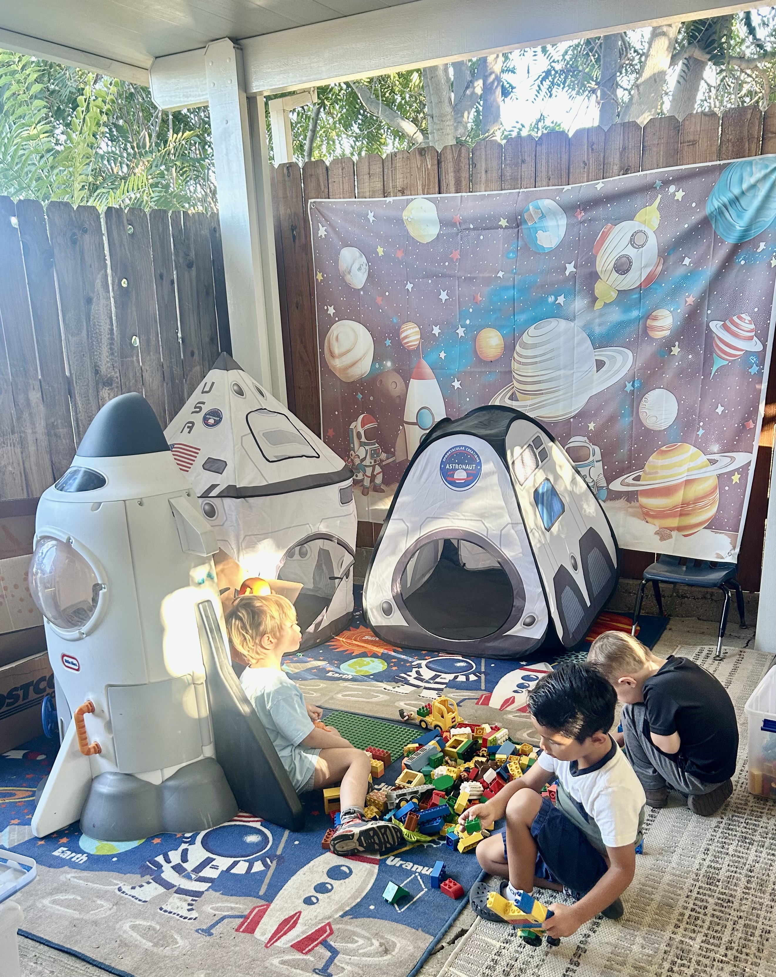 Children playing with building blocks between rocket ships and astronaut tents in the backyard play area