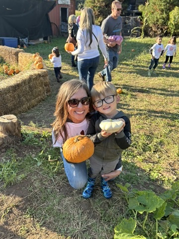 Miss Robin kneeling beside a preschooler holding a white pumpkin at the pumpkin patch