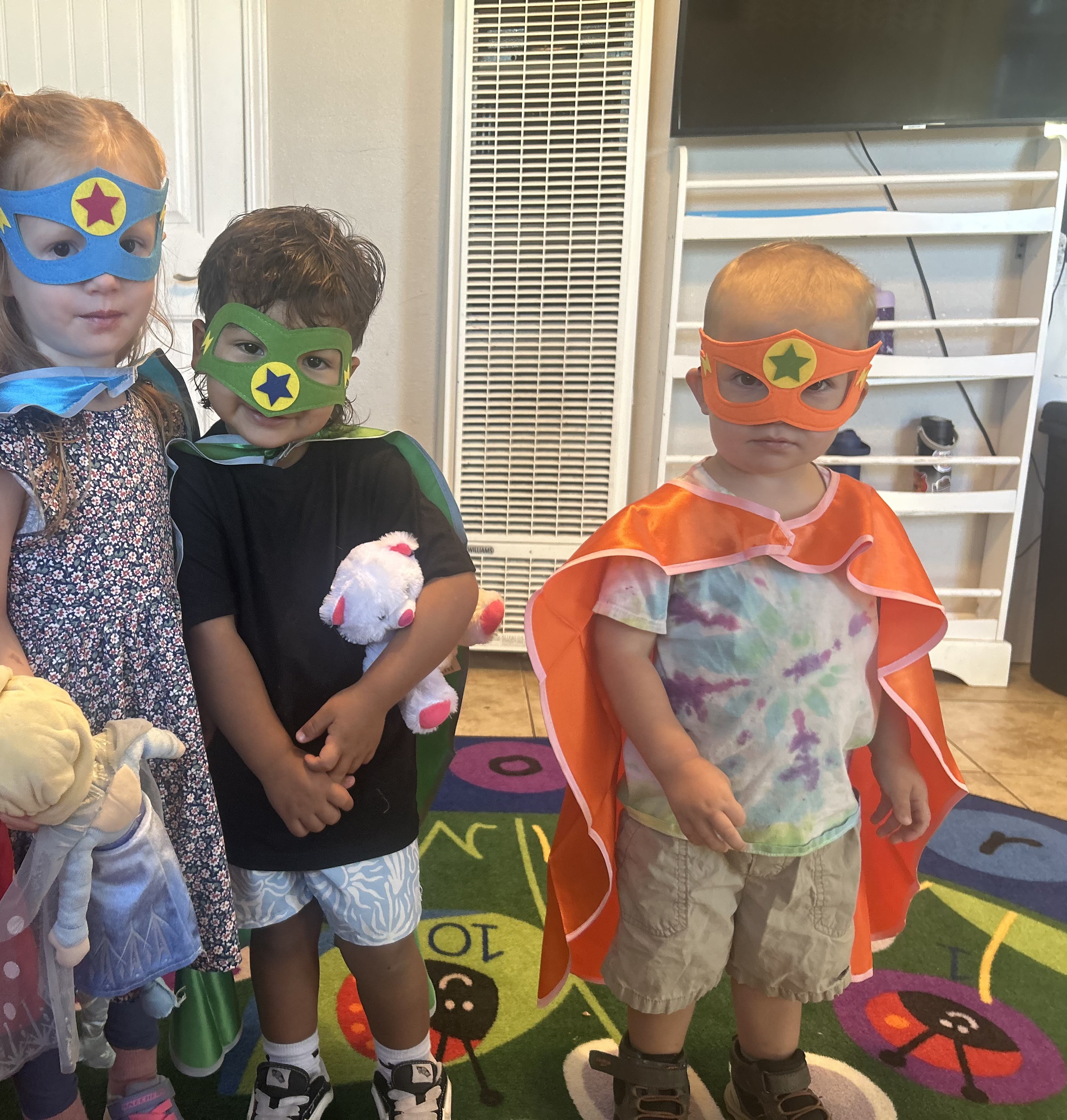 Four preschoolers wearing superhero masks and capes standing together in the classroom