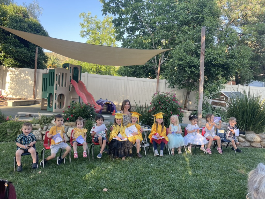 Miss Robin and the preschoolers seated in chairs on the backyard lawn for the graduation ceremony
